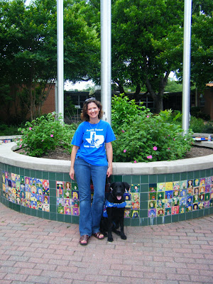 I'm standing with a black lab in working jacket, in front of a colorful round tiled planter with school flagpoles in the middle