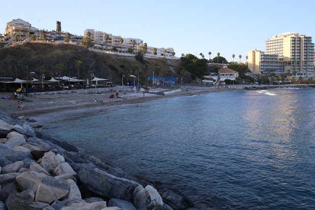 Playa con el agua del mar, escolleras de rocas a un lado y un acantilado con vegetación y edificios altos a su espalda.