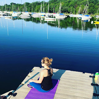 Woman sitting on yoga mat on a dock with sailboats in the background.
