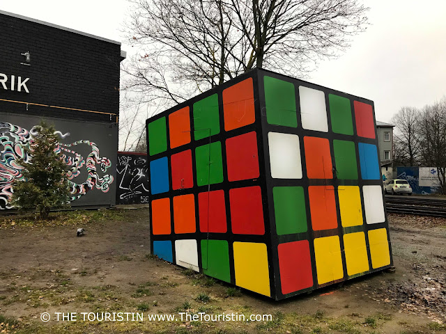 A giant-sized Rubik's cube on a muddy square in front of a two storey industrial stye warehouse, under a light grey winter sky.
