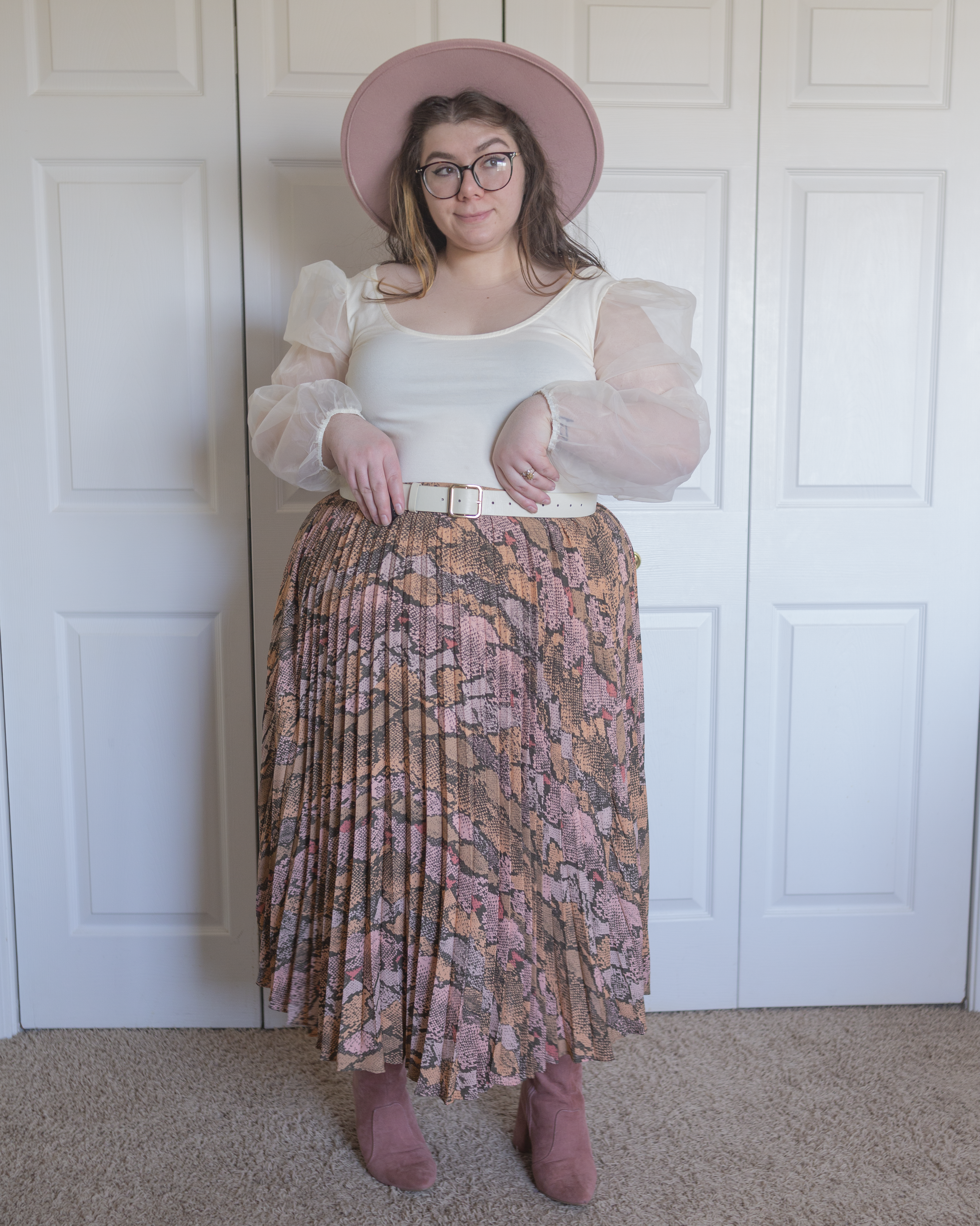 An outfit consisting of a pastel pink wide brim fedora, a cream colored wide scoop neck blouse with long sheer balloon sleeves tucked into a pastel pink, orange, brown and gray snake print accordion pleated midi skirt and pastel pink suede midi boots.