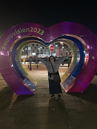 Me, with outstretched arms and joy in my heart, poses inside a massive heart sculpture. It says EUROVISION 2023 on the side of it. It's night time.