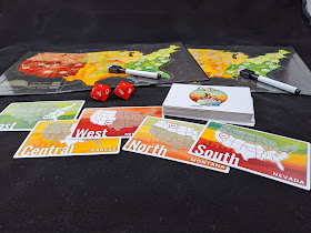 Two fold-out game boards with a map of the continental USA with dry-erase markers, laid out behind a stack of cards and two large ten-sided dice. Five cards have been turned over: one shows Nevada with the southern half of the country highlighted, another shows Montana in the highlighted North half, one shows New Mexico in the highlighted West region, one shows Kansas in the Central section, and the last one shows Mississippi in the highlighted East region.