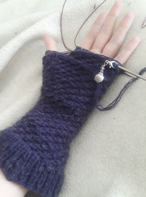 a dark purple mitten in progress, knit in a spiralling star stitch pattern.   The working yarn is 4 strands of very fine yarn. There is a silver stitch marker with a shell charm hooked over one of the needles.