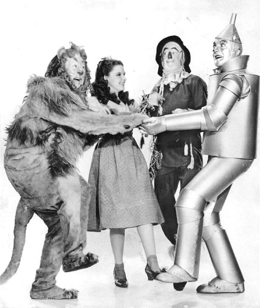 black-and-white publicity still of (left to right) a man in a lion costume, a girl in a pretty dress, a man dressed as a scarecrow, and a man made of tin. All seem to be animated and laughing.