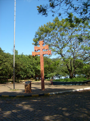 Foto em plano geral e na vertical mostrando a Cruz Missioneira, ao fundo algumas árvores.
