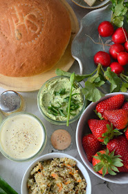 Die Zutaten für einen bunten Veggie-Weihkorb zum Osterfest liegen auf einem grauen Untergrund: Osterbrot, Radieschen, frische Aufstriche, gefärbte Eier, Bärlauch und Erdbeeren.