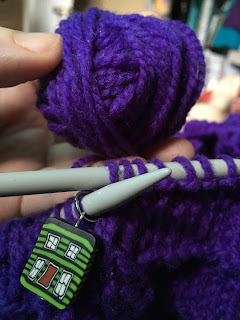 A close up photo of a small hand wound ball of purple wool being held just above the project in progress on its circular knitting needle. There is a bright green house stitch marker on the left needle along with the stitches waiting to be cast-off.