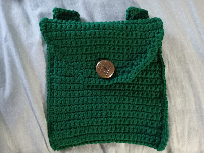 A green crocheted pouch with a brass button. The pouch is slightly too long to be square and has a slightly tapered flap that is held down by the brass button, which has a pattern of intersecting lines on it.