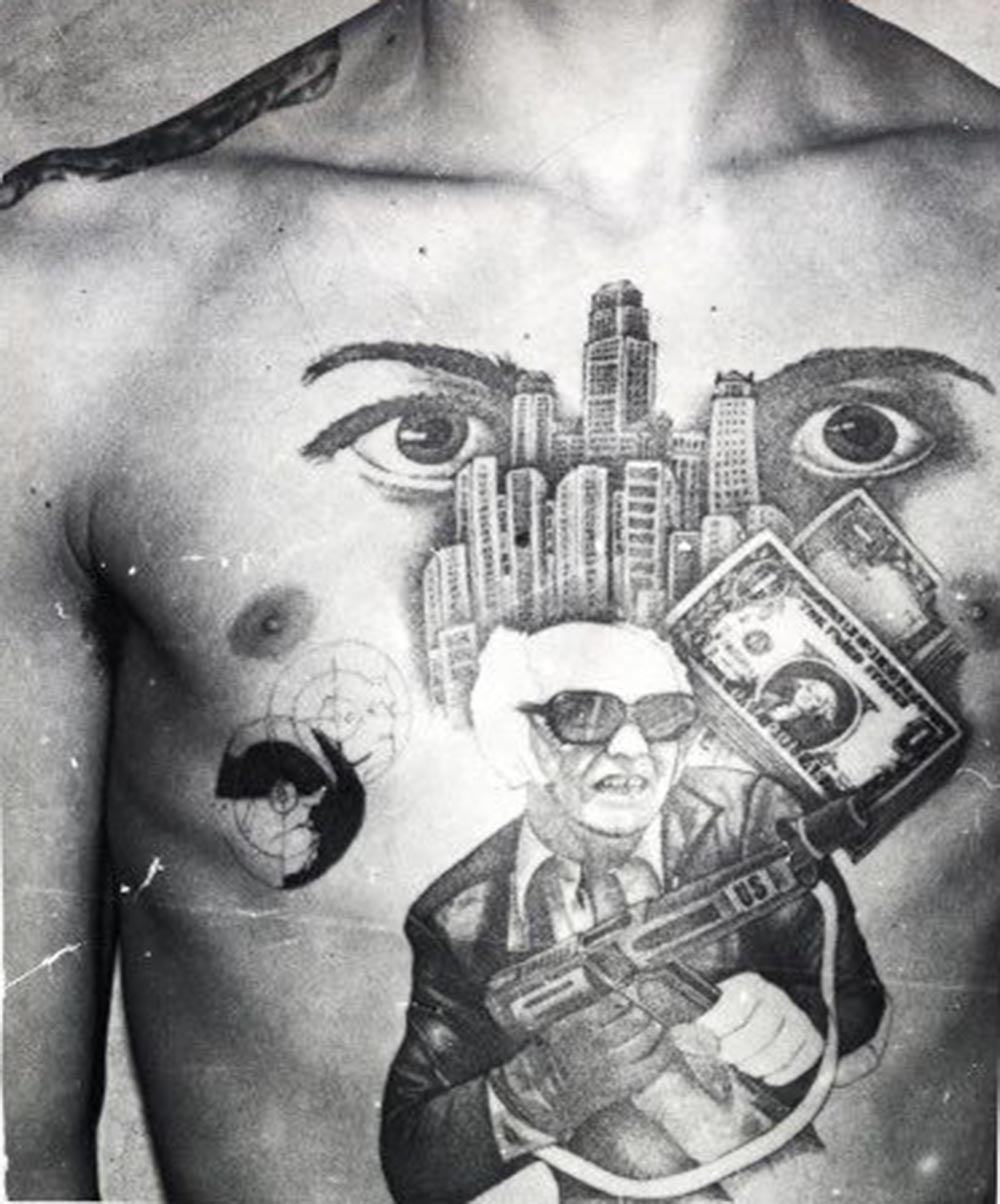 The dollar bills, skyscrapers and machine gun with the initials 'US' stamped on it convey this inmate's love for the American mafia-like lifestyle. The eyes signify 'I'm watching over you' (meaning the other inmates in the prison or camp).