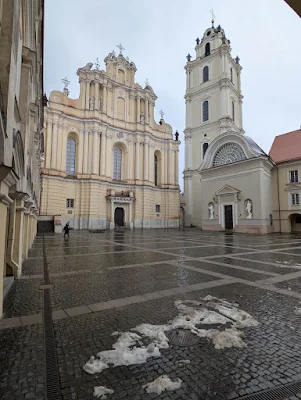 A wide view of a large, wet cobblestone square in front of two grand Baroque-style buildings. The building on the left is a church with an ornate facade, and the building on the right is a tall bell tower with an arched entrance. Patches of dirty snow are on the cobblestones.