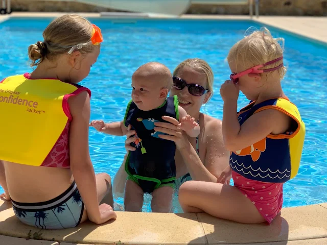 2 girls sitting on the edge of the pool in konfidence swim jackets and a baby being held up to them by the mum. The baby wears the Konfidence Babywarma on a sunny day in France