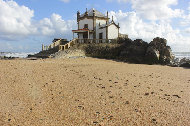 Wzniesiona na skale, sześcioboczna kaplica o pobielonych ścianach. U jej stóp leży piaszczysta plaża. W tle widoczny jest rozfalowany ocean.