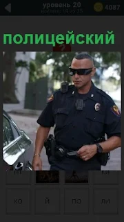 Около машины стоит полицейский в форме и темных очках и с рацией на плече 
