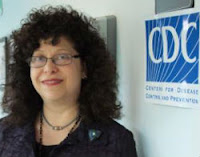 a plump middle-aged white woman with fluffy permed black hair and wire rimmed glasses stands next to a sign for the Centers for Disease Control