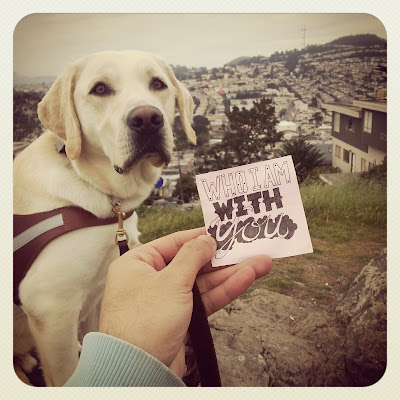 JR holds up one of his unique typography notes on a post-it that reads "Who I am with you" - his guide dog Griff sits looking at the camera and behind him overlooks San Francisco.
