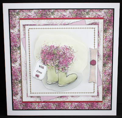 The image is a photo of a multi-layered and crafted pink and white square floral card, featuring a watercolour drawing of pink flowers growing out of green wellies in the centre.