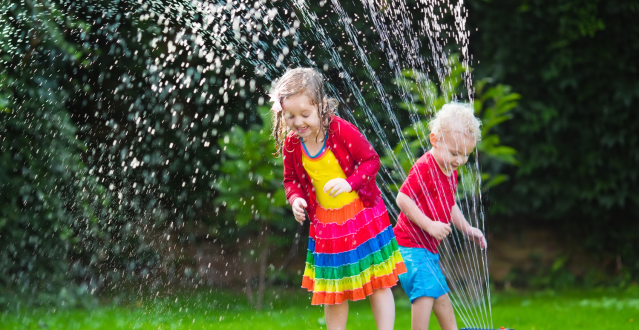 Girl and boy playfully dash through garden sprinkler, laughter echoing in the air.