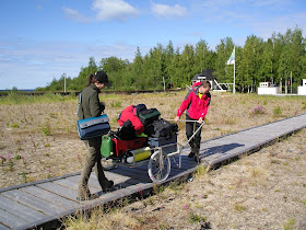 Kaksi henkilöä kärrää maitokärryillä sukellusvarusteita pitkoksia pitkin hiekkarannalla