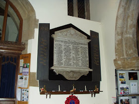 A white painted church wall, with a small window placed very high up.  The memorial tablet is decorated top and bottom with carving and there are four columns of names upon it.  On either side are dark slate strips with gold painted names for the 1939-45 war.  A shelf below carries candles and poppies.