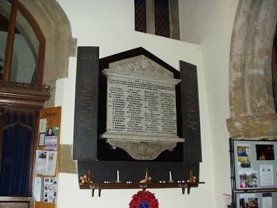 A white painted church wall, with a small window placed very high up.  The memorial tablet is decorated top and bottom with carving and there are four columns of names upon it.  On either side are dark slate strips with gold painted names for the 1939-45 war.  A shelf below carries candles and poppies.