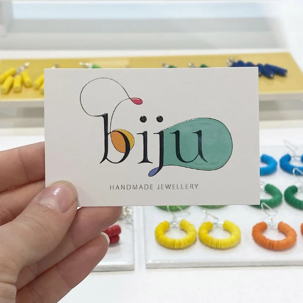 hand holding a Biju Jewellery business card with paper earrings displayed in background.