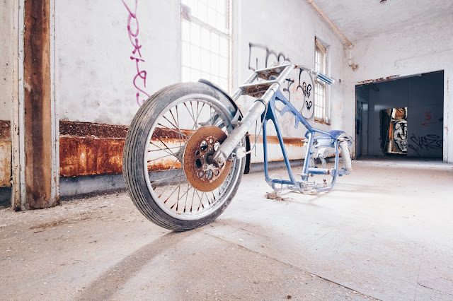 A sunlit, abandoned room with a rusted, overturned bicycle in the foreground, and graffiti on the walls.