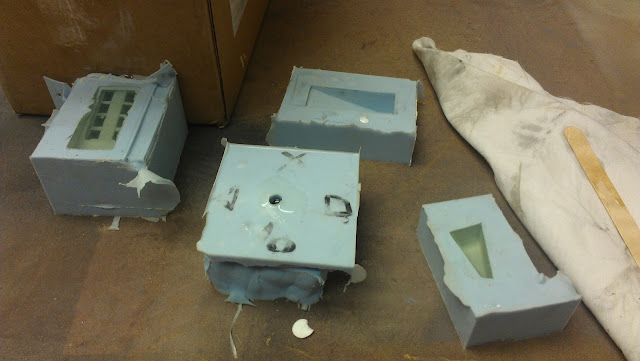 On a grayish brown table sits a cardboard box and a rag with a stick on it.  There are also four blue rubbery molds, each one a rectangular shape.  In each mold is a different shape though; a couple wedges of different sizes, a rectangle with eight perpendicular rectangles sticking out of it, and another with a blue rubber lid on it with a whole and a triangle, circle, square and x marking each side of the lid.  All but one wedge are filled with a clear, yellowish liquid that will soon solidify and turn hard and white to become the model. 