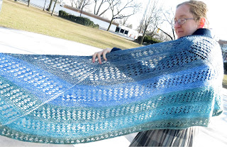 A woman wearing a garter stitch and lace crescent shawl. The shawl is striped in a blue-green gradient. One edge of the shawl is out of frame, with the other end wrapped around her shoulder, with the wingspan held open.