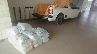 A imagem mostra uma pickup branca estacionada no salão de entrada da Arena Pernambuco várias sacas de adubo empilhadas no fundo. As paredes do salão são feitas de concreto. O chão é feito de concreto. Não há pessoas na imagem.