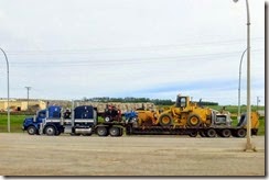 Two trucks with very large equipment.  behind are trimmed logs piled high