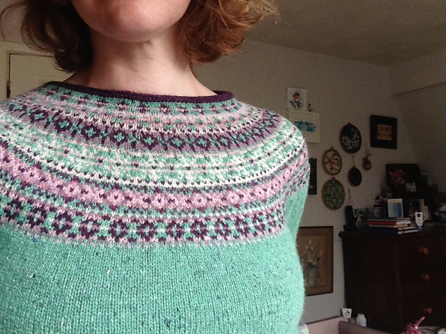 A woman with untidy bobbed hair models the yoke of her hand-knit jumper. The yoke has subtle spot and diamond patterns knit in pink, purples, green and white. On the wall behind her are small embroidered hoops with floral designs. There is also an old dark chest of drawers with a stack of books on its surface.