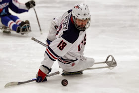 picture of josh sweeney on  the ice playing sled hockey with the puck and two shorter hockey sticks