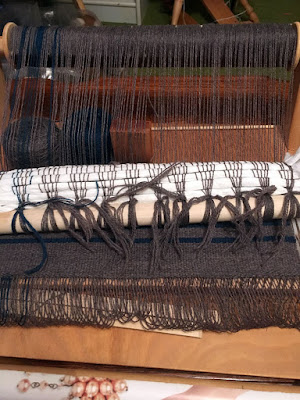 A small, natural wood rigid heddle loom with grey warp threads and three uneven stripes of deep teal at the left edge wound on the back beam. The warp threads are knotted onto a pale wooden dowel at the near edge, with six rows of scrunched toilet paper woven in just above the knots, and a small piece of mostly grey weft-faced fabric showing folded underneath the dowel. Two narrow teal stripes show across the fabric as well, and one cake each of grey and teal yarn peep through the warp threads at far left.