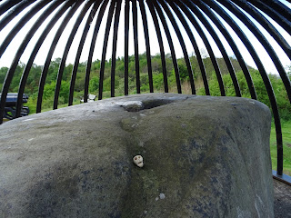A photo of a small, ceramic skull (Skulferatu 74) lying in a hollow in the Beheading Stone on Mote Hill in Stirling.  In the background are the iron bars of the cage in which the stone is kept in.  Photograph by Kevin Nosferatu for the Skulferatu Project.