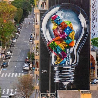 Foto em perspectiva e do alto. À esquerda, a rua Oscar Freire em São Paulo, ladeada por copas de árvores e carros estacionados À direita, a parede lateral de um prédio de seis pavimentos com um mural de 30 metros de altura,  pintado pelo artista Eduardo Kobra. Sobre fundo preto, uma lâmpada incandescente.  Dentro, a pintura cubista, com formas fragmentadas e geométricas coloridas, remete à escultura em bronze de Auguste Rodin intitulada “O Pensador” um homem nu,  sentado sobre uma pedra com os pés apoiados na base da pedra. O corpo levemente curvado tem músculos bem delineados; está com o punho esquerdo apoiado sobre o joelho esquerdo com a mão pendente; o tronco em leve torção à esquerda posiciona o cotovelo direito apoiado sobre a coxa esquerda com o dorso da mão voltado para cima e para dentro; a boca está apoiada no dorso da mão que sustenta a cabeça. Sua expressão é de profunda meditação e questionamento). Andaimes e cordas ainda estão visíveis na base do prédio. Kobra está no andaime esquerdo e finaliza a obra.   Detalhe: Texto do post: SP ganhou um novo mural do artista @kobrastreetart!  O nome do novo trabalho, que fica na Rua Oscar Freire, nos Jardins, é Seja Luz, e o objetivo, segundo o artista, é fazer com que repensemos, nestes tempos de pandemia, nossos valores, conceitos e princípios.  E também, "que possamos apresentar nossas soluções ao mundo, que nunca esteve tão necessitado de respostas para questões cruciais, que vão da fome à violência, liberdades e tolerância ao outro. Se há trevas, que sejamos luz."