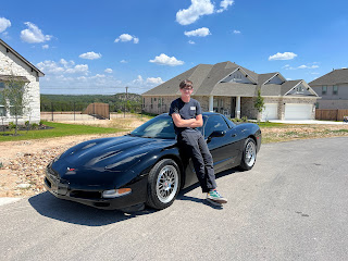 TJ, a tall teenager, leans against his black 1997 Corvette that is parked in the street in front of houses and blue sky.