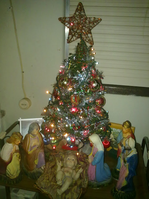 Uma foto com uma árvore de Natal decorada com enfeites coloridos e piscas, ao lado de um presépio com figuras de Maria, José e o menino Jesus