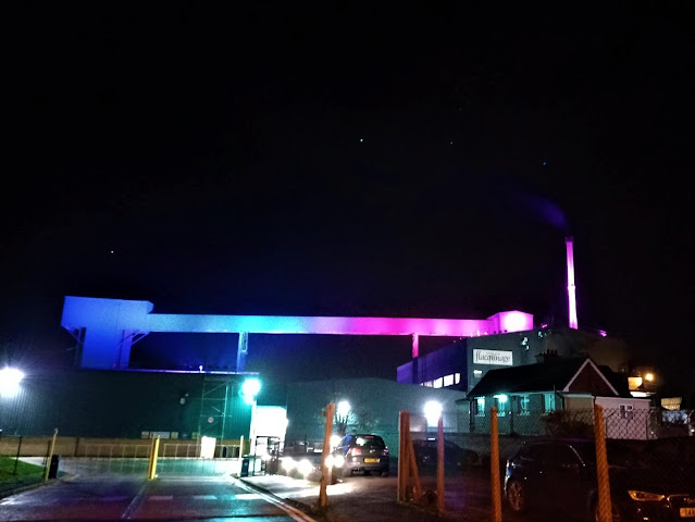 Part of the Stoelzle Flaconnage glassworks lit up in blue, green and pink against the dark November night's sky. Steam is coming out of the large chimney.