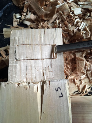 A photo from above showing the end of a beam and the tenon protruding from it, with the smaller rectangular mortise cut around the edges and a narrow chisel set to chip out the first layer of wood.