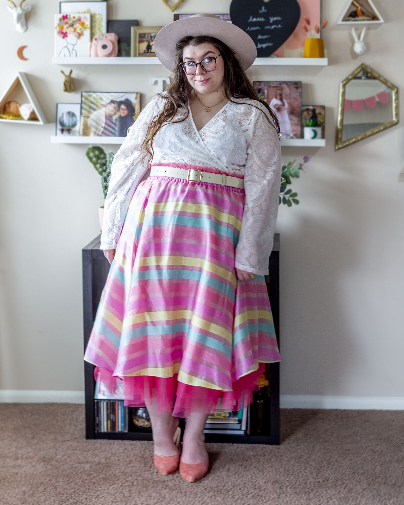 An outfit consisting of a beige and pink wide brim porkpie hat, a white lace wrap top with angel sleeves tucked into a pastel pink with pastel pink, lemon yellow and sky blue horizontal stripes with hot pink tulle exposed at the bottom hem, and pastel pink heeled clogs.