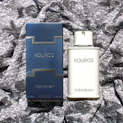 The white, opaque bottle and blue box of Kouros by Yves Saint Laurent lying flat on a black and white, textured, crinkled sweater