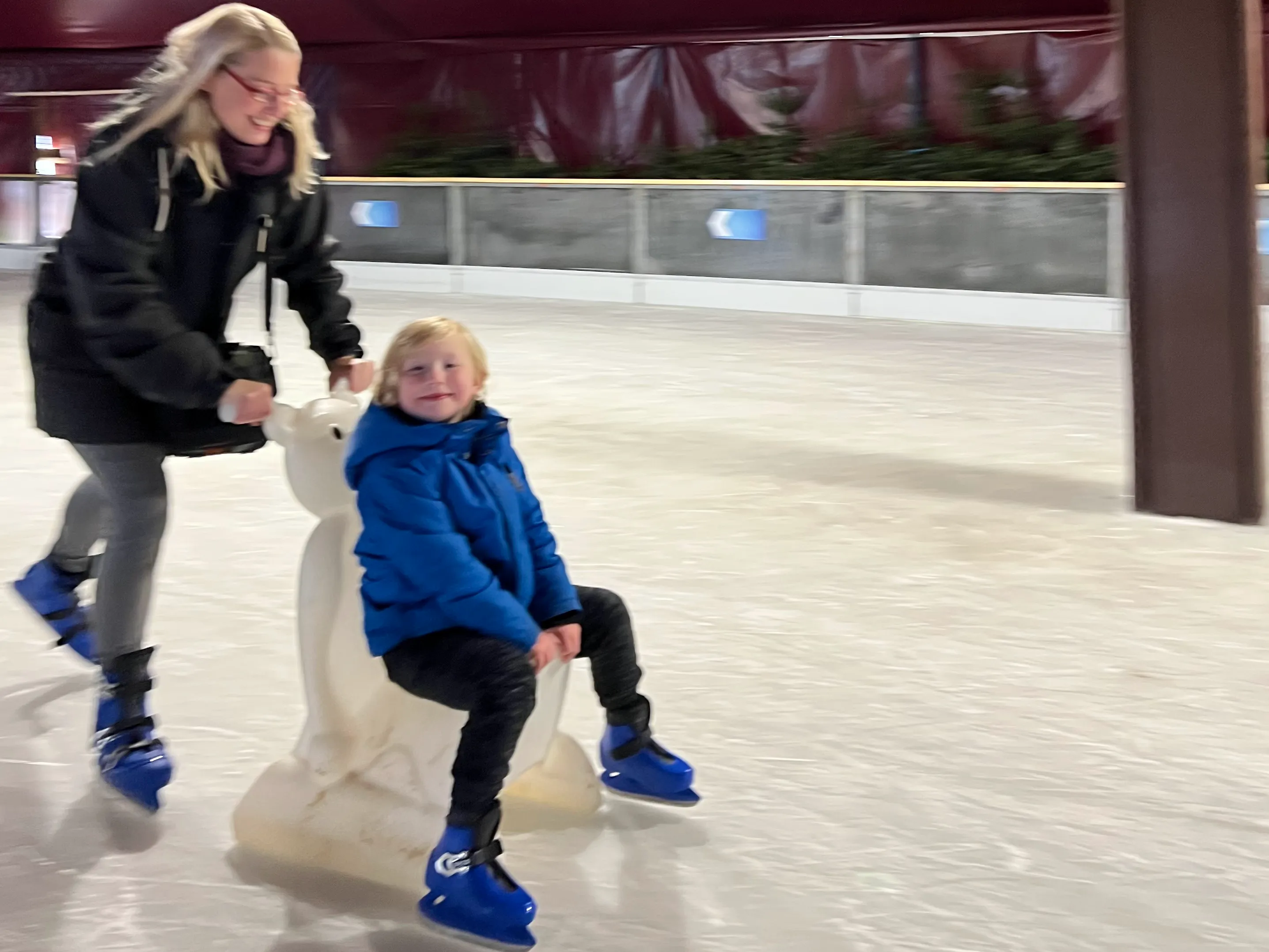 An adult pushing a child around a seasonal ice rink on a skate aid