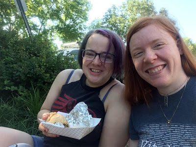 Two young women smiling at the camera. One has glasses and short purple-dyed hair, and the other has long red hair. They're sharing tamales in a paper boat while sitting in the shade.