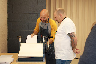 two people stand at a printing press, feeding the plate and paper into the press