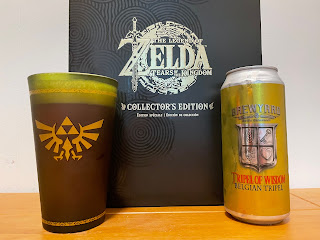 Tripel of Wisdom poured into a green pint glass featuring the Triforce beside the Collector's Edition of Tears of the Kingdom and the beer's can.