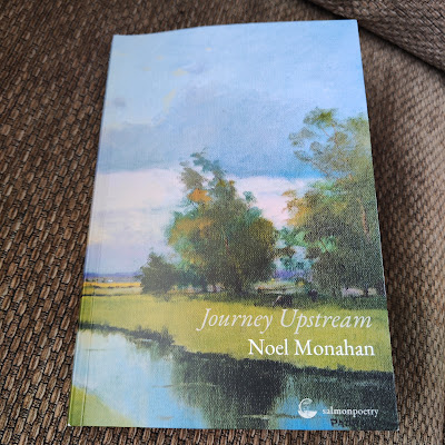 a book sits on a couch, the title is Journey Upstream, the author is Noel Monahan, the picture is of a rural painting with river and fields and trees and sky