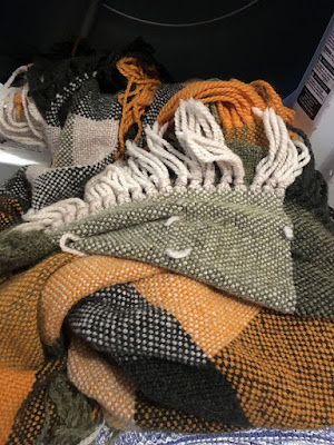 A crumpled and twisted length of plain-woven plaid cloth, in white, light and dark green, and marigold, with long fringe, hanging over the edge of an open dryer. A few fluffy tails of yarn protrude from the surface of the cloth.