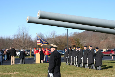 Men in military uniform gathered for tribute ceremony under two long gray battleship guns. A podium flanked by flags is in the background.