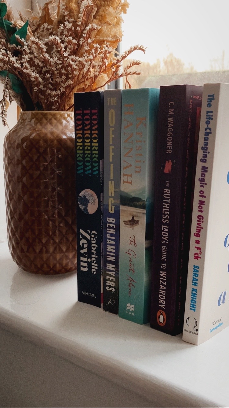 a line of books on a windowsill next to a vase of dried flowers